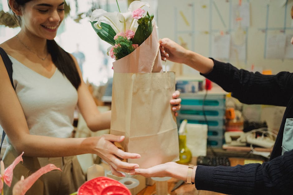 A woman with long dark hair and a white sleeveless top is smiling as she accepts a floral bouquet from another person at a florist shop. The bouquet, wrapped in pink paper and enclosed in a beige pape