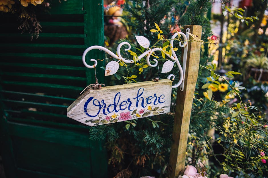 A rustic wooden sign with the words 'Order here' painted in blue cursive, hanging from a white wrought iron bracket adorned with decorative swirls and small vine-like leaves. The sign is nestled among