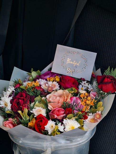 A vibrant floral bouquet arranged with a variety of fresh flowers including red and pink roses, white daisies, peach-colored blooms, yellow and purple accents, and green foliage. The bouquet is wrappe