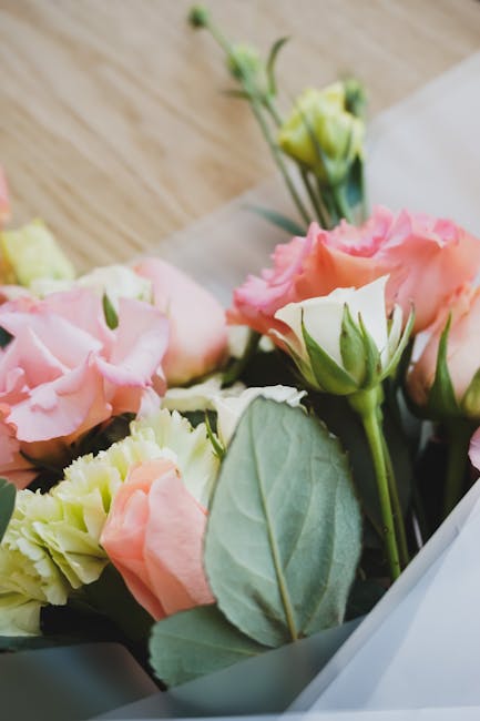 A close-up of a floral arrangement featuring soft pink and creamy white roses and lisianthus, with lush green eucalyptus leaves, wrapped in light grey paper. The bouquet is resting on a wooden surface